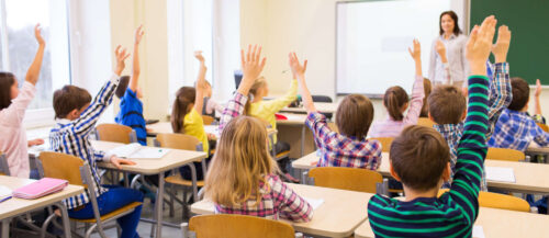 group of school kids raising hands in classroom